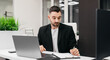 © wedmoments.stock - Businessman in black blazer is working at a modern office desk with a laptop and notepad, surrounded by greenery and contemporary decor during a focused work session