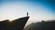 © CreativeIMGIdeas - Silhouette of a man standing on the edge of a steep cliff overlooking mountain peaks at sunrise, symbolizing freedom, challenge, and success.