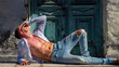© Volodymyr - Shirtless man relaxing in sunlight with open shirt and denim jeans. Confident male model posing outdoors against a vintage door background. Sensual lifestyle portrait of a fit man enjoying summer.