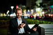 © Volodymyr - Man in business suit talking on phone at night. Business man making a call on night street. Business man with a tablet and phone outdoors. Businessman sitting on bench in night street. Night business.