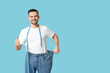 © Pixel-Shot - Handsome sporty young man in loose jeans with measuring tape showing thumb-up gesture on blue background. Weight loss concept