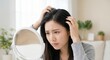 © Gayan - Concerned young Asian woman examining hair loss and scalp health in a vanity mirror indoors, pensive adult female worrying about thinning hair and beauty care in a bright room.