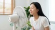 © Gayan - A relaxed young East Asian woman in a white t-shirt sitting on a sofa and cooling off in front of a white electric fan in a bright modern living room during a hot summer day indoors.