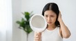 © Gayan - Concerned Southeast Asian adult woman examining her hair and scalp in a mirror indoors during the morning, hair care health and hair loss concept, worried expression, natural light.