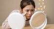 © Gayan - Worried young Asian woman looking in a round white makeup mirror and pointing at dark circles under her eyes, concerned about skincare and aging in a bright indoor room with soft lighting.