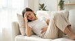 © Gayan - Young Southeast Asian woman lying on a white sofa indoors, holding her forehead and stomach in pain, suffering from menstrual cramps or a stomach ache, lifestyle healthcare concept.
