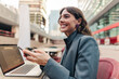 © Westend61 - Businesswoman smiling holding phone with laptop at public space in Madrid