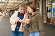 © Iryna - Two friends look at a phone while standing at a train station in the afternoon
