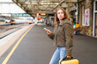 © Iryna - Waiting at the train station while holding a phone and a suitcase in the afternoon