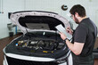 © pressmaster - Caucasian young adult man inspecting engine compartment of car while holding clipboard and writing notes in auto repair shop, standing beside open hood and focusing on checklist