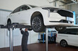 © pressmaster - Black man mechanic working under elevated sedan in auto repair shop, inspecting vehicle underside, wearing uniform, standing on workshop floor, focusing on maintenance task