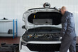 © pressmaster - Middle aged Caucasian man inspecting engine compartment of modern car in auto repair shop, standing beside open hood and wearing work gloves, focusing on vehicle maintenance