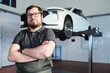 © pressmaster - Portrait of Caucasian middle aged man with beard and glasses standing with arms crossed in auto repair shop, car lifted on hydraulic platform in background, looking at camera