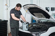 © pressmaster - Caucasian young adult man wearing glasses preparing to pour motor oil into car engine while standing in auto repair shop with hood open, focusing on maintenance task