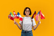 © Prostock-studio - Happy lady showing bunch of diverse flags cheerfully smiling at camera over yellow studio background, excited female student recommending foreign language studying school