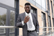 © Prostock-studio - Happy black businessman with smartphone in hand reading good message and chatting about deal, standing outdoors near office center