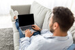© Prostock-studio - Advertisement concept. Young guy using modern laptop with blank screen, sitting on sofa, mockup. Millennial man reclining on couch and using notebook with blank display, over shoulder shot