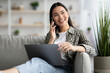 © Prostock-studio - Beautiful young asian woman having phone conversation, sitting on couch in living room and using laptop, looking aside, copy space. Cheerful lady resting at home, websurfing and talking on smartphone