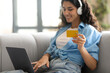 © Prostock-studio - Pretty millennial woman with laptop and headphones showing credit card at camera, sitting on couch at home, selective focus. Young lady shopping online, using remote banking service, copy space