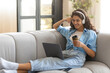 © Prostock-studio - Happy young brunette woman in headphones having online video conference on laptop, drinking coffee on couch at home, free space. Millennial lady studying or working on web at living room