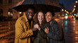 © Николай Срибяник - A joyful group of three diverse friends laughing together under one umbrella on a rainy city street at night.
