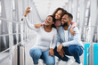 © Prostock-studio - Ready For Adventures. Portrait Of Cheerful Young Black Family Taking Selfie With Smartphone In Airport Terminal Or On Railway Station, Tourists Posing At Camera Together, Blurrred Background