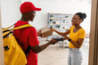 © Prostock-studio - Door Delivery. Happy Black Customer Lady Receiving And Paying For Package Box With Phone Taking Parcel From Courier Guy In Red Uniform Standing In Doorway At Home. Modern Shopping Concept