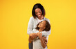 © Prostock-studio - Mother's love and care. Happy young african american mom embracing her cute little daughter from the back, standing over yellow background, free space