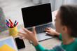 © Prostock-studio - Over the shoulder view of girl sitting at desk, making video call with teacher or class group using laptop with black blank screen, waving hand and talking, selective focus, mock up, free space