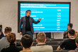 © Anton Gvozdikov - Speaker presents data to attentive audience modern conference room. Blue screen displays statistics and graphs. Professional meeting atmosphere.
