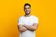 © Prostock-studio - Positive handsome middle-eastern guy in white t-shirt posing on yellow studio background, copy space. Cheerful young good-looking arab man with arm crossed on chest smiling at camera