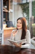 © bongkarn - Pretty asian woman holding notebook thinking with pen under chin sits at wooden table outside cafe.