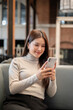 © bongkarn - Pretty asian woman holding and looking at phone while sitting leaning on sofa in coffeehouse or cafe