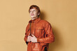 © SHOTPRIME STUDIO - Young man with red hair wearing brown leather jacket standing against beige background. Portrait of confident male model looking up with smile and casual pose in studio light.