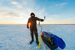 © True Pixel Art - A bundled angler stands on a frozen lake at sunrise, gripping a hand auger. A sled with gear, folding chair, and a bright blue tarp rests nearby in cold dawn light.