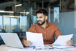 © Liubomir - Young indian businessman at a modern office desk, smiling while reviewing reports and laptop, planning finances and strategy with confident, focused professionalism and productivity