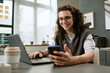© AnnaStills - Young adult Caucasian man sitting at desk using smartphone and laptop, smiling while working in modern office setting, takeaway coffee cup and computer screen in background