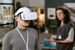 © AnnaStills - Young adult Caucasian man wearing virtual reality headset standing in modern office, while young adult man with long curly hair and glasses holding digital tablet in background observing