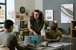 © AnnaStills - Young adult Caucasian man standing and gesturing while talking to young adult Caucasian man and young adult Black man, sitting at desk using laptops in modern office workspace