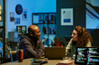 © AnnaStills - Young adult Black man and young adult Caucasian man sitting at desk discussing project in modern office, both focused on conversation with laptops and computer monitors displaying code