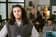 © AnnaStills - Portrait of Caucasian teenager boy with long curly hair standing in modern office looking at camera, other young men working at computers in background