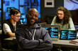 © AnnaStills - Portrait of young Black man smiling with arms crossed in front of computer screens displaying code while young adult Caucasian men working behind