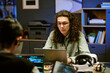 © AnnaStills - Young adult Caucasian man with long curly hair working on laptop at desk surrounded by computers and virtual reality headset, focusing on screen in modern office setting