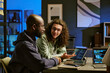 © AnnaStills - Young adult Caucasian man with long curly hair discussing programming code with young Black man, while sitting at desk with laptops in modern office environment