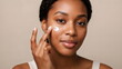 © xuxu - An African American woman is applying face cream to her problem skin with acne. Daily skincare and cosmetic procedures. Skin care, cosmetics, and moisturizing. On a beige background.