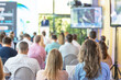 © wellphoto - Business conference audience attending keynote presentation in modern seminar hall, corporate event training with speaker on stage and screen