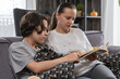 © dimetradim - Mother and teenage son sitting on sofa reading book together at home. Boy points at page while woman explains text in living room interior. Family education and bonding during leisure time.