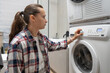 © dimetradim - Woman starting laundry cycle by turning program selector dial on washing machine panel. Housewife choosing wash mode in bathroom with stacked dryer appliance for domestic cleaning routine.