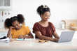 © Prostock-studio - Happy african american school girl teenager doing homework, sitting at table by her cheerful working mother, kitchen interior. Black lady freelancer working from home, using laptop