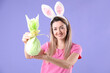 © New Africa - Easter celebration. Smiling woman in headband with bunny ears holding wrapped gift on violet background